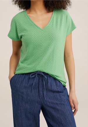 Woman wearing green short-sleeve V-neck top with subtle chevron pattern and blue drawstring pants, hands in pockets, neutral background.