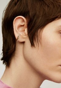 Aro de oro con un diseño elegante, que presenta pequeñas piedras preciosas transparentes en el borde exterior, lucido por un modelo de cabello corto.