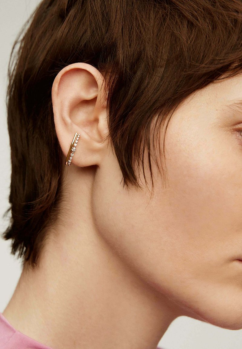 Aro de oro con un diseño elegante, que presenta pequeñas piedras preciosas transparentes en el borde exterior, lucido por un modelo de cabello corto.