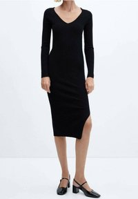 Abito a maglia nero a coste con maniche lunghe, scollatura a V, lunghezza a metà polpaccio, spacco laterale, abbinato a scarpe nere con tacco a blocco.