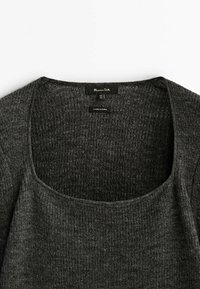 Pull en maille gris charbon avec un large col rond, une texture côtelée, et une étiquette portant le nom de la marque ainsi que les informations d'entretien.