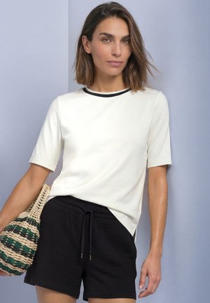 Kvinde med skulderlangt hår har en hvid top med korte ærmer og sorte shorts på, mens hun holder en flettet taske med grønne og beige striber.