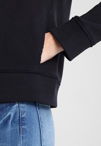 Felpa blu navy con dettagli di polsini e orlo a coste, dotata di tasche laterali; il tessuto ha una consistenza morbida e bordi leggermente sfilacciati.