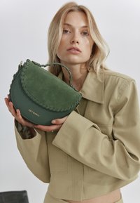 Sac à main en cuir vert avec une forme courbée, une surface texturée et des accents tressés. Doté d'une poignée supérieure et d'un détail logo doré sur le devant.