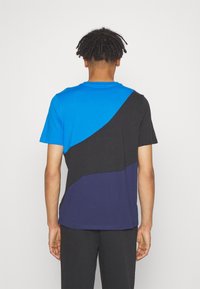 Tričko s krátkým rukávem, které má geometrický design v barevném bloku v odstínech modré, černé a tmavě námořnické, s hladkým bavlněným povrchem.