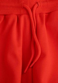 Czerwone dresowe spodnie z rypsowym paskiem w talii i plecionymi sznurkami. Materiał ma gładką fakturę i matowe wykończenie.