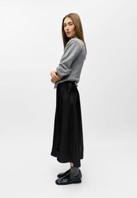 Grå strikket sweater og sorte satin midi-nederdele med en knyttet talje. Modellen har grå strømper og sorte glansfulde sko på, mens hun står foran en hvid baggrund.
