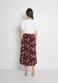 Weißes Kurzarmoberteil kombiniert mit einem langen bordeauxfarbenen Rock, der mit einem floralen Muster in Weiß und Schwarz verziert ist. Dazu werden weiße High-Heels getragen.