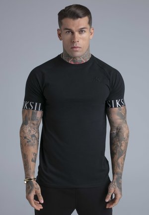 Mees, kellel on kaelal ja käel tätoveeringud, kannab musta formis T-särki, mille varrukatel on kiri "SIKSILK", seisab tavalise halli tausta ees.