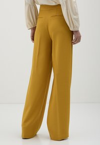 Persona che indossa pantaloni a gamba larga giallo senape e una blusa beige a maniche lunghe, in piedi davanti a uno sfondo chiaro e uniforme.