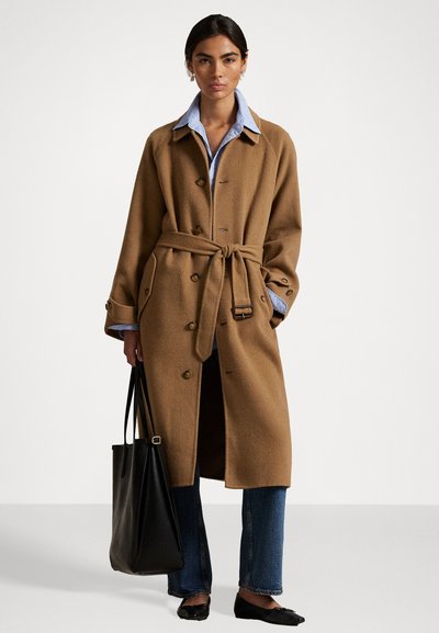 Brown wool trench coat with a belted waist, large buttons, and side pockets. Worn over a light blue collared shirt and dark jeans. Black tote bag.