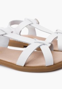 White faux leather sandals with crisscross straps, flat tan sole, and textured insole. Close-up view highlighting strap detail and color contrast.