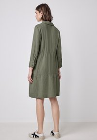 Abito verde oliva con scollatura a collare, maniche lunghe, design a strati e tessuto leggero. Indossato con calzature casual.