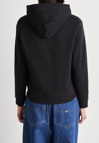 Černá bavlněná mikina s kapucí na stahování, ribbed manžety a lem, v kombinaci s modrými širokými džíny se dvěma zadními kapsami.