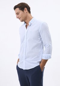 Camisa de manga larga a rayas en azul claro y blanco con cuello abotonado, que presenta un pequeño logotipo bordado y puños enrollados.