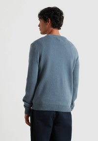 Mężczyzna z kręconymi ciemnymi włosami, ubrany w jasnoniebieski sweter z dzianiny i ciemne spodnie, lekko obrócony w lewo na jednolitym jasnym tle.