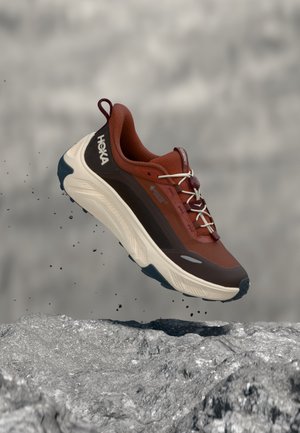 Chaussure de course HOKA marron et beige avec des accents noirs en suspension au-dessus d'un terrain rocheux avec des particules de poussière en dessous.