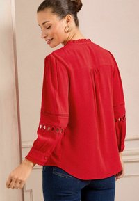 Blusa rossa con colletto arricciato, maniche lunghe con dettagli decorativi, piega sulla schiena e una texture liscia. Abbinata a jeans blu.
