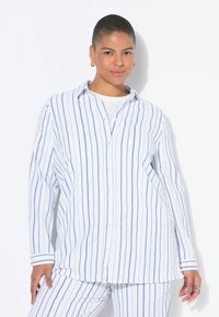 Camicia a righe in tessuto bianco e azzurro chiaro, con bottoni, maniche lunghe, due tasche frontali, vestibilità rilassata, indossata con pantaloni a righe coordinati.