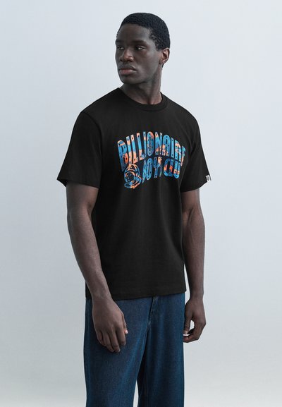 Νεαρός άνδρας που φοράει μαύρο T-shirt με πολύχρωμο κείμενο και γραφικό «Billionaire Boys Club», συνδυασμένο με μπλε τζιν, στέκεται μπροστά σε απλή φόντο.