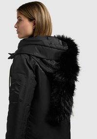 Sort pufferjakke med hætte, som har en trim i falsk pels. Jakken har en glat overflade og en lynlåslomme i siden.