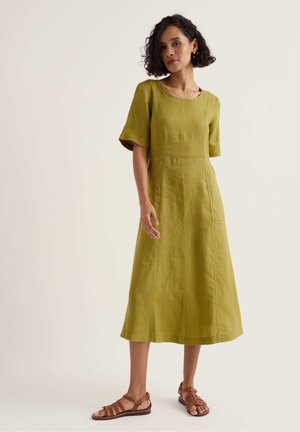 Žena s kovrčavom kosom, odjevena u haljinu maslinasto zelene boje srednje dužine s kratkim rukavima i smeđim sandalama s remenčićima, stoji na jednostavnoj pozadini.