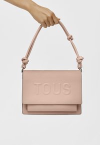 Różowa skórzana torba na ramię z wytłoczonym logo "TOUS", płaski design oraz węzełkowy pasek na ramię. Gładka tekstura i prostokątny kształt.