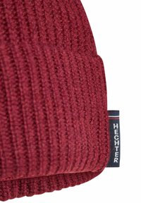 Rode gebreide muts met geribbelde structuur. Voorzien van een ingestikte label met de tekst "HECHTER" in wit op een blauwe achtergrond.