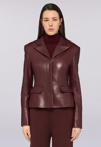 Giacca di pelle borgogna con colletto a scialle, tasche frontali e design aderente, abbinata a un dolcevita coordinato. Texture liscia.