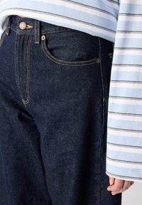 Jeans in denim blu scuro con cuciture arancioni, dotati di una tasca frontale e chiusura con bottone. Camicia a righe blu chiaro parzialmente visibile.