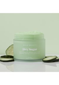 Eksfolierende kroppsskrubb i en gjennomsiktig, lys grønn krukke med en teksturert lokk. Akkompagnert av agurkskiver, plassert mot en lys grønn bakgrunn.