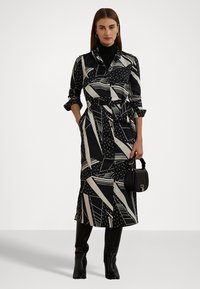 Svart och vit mönstrad klänning med geometriska former, midjebälte, krage och långa ärmar, kombinerad med en texturerad handväska.
