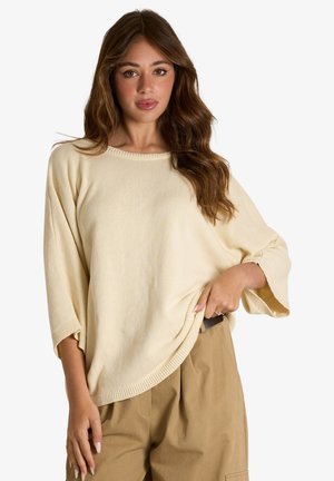 Donna con capelli castani lunghi che indossa un maglione crema largo con maniche ampie e pantaloni beige a vita alta su sfondo bianco.