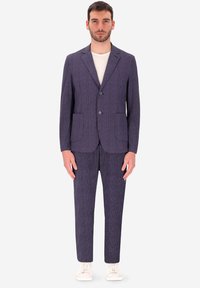 Uomo in piedi che indossa un abito blu navy a righe con due bottoni, abbinato a una maglietta bianca a girocollo e sneakers bianche.