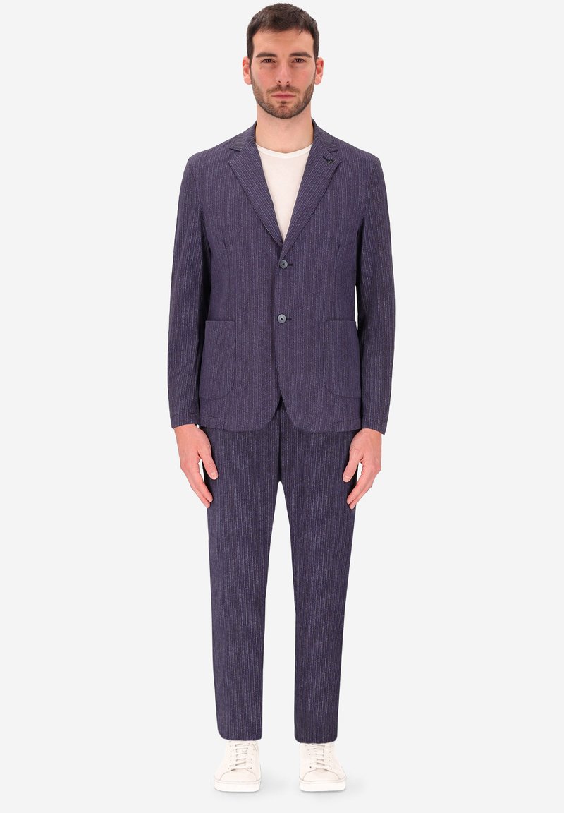 Uomo in piedi che indossa un abito blu navy a righe con due bottoni, abbinato a una maglietta bianca a girocollo e sneakers bianche.