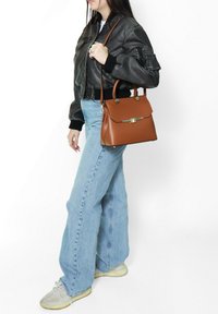 Borsa a tracolla in pelle marrone con hardware dorato, design strutturato, doppi manici e tracolla staccabile; il modello indossa una giacca bomber nera e jeans.