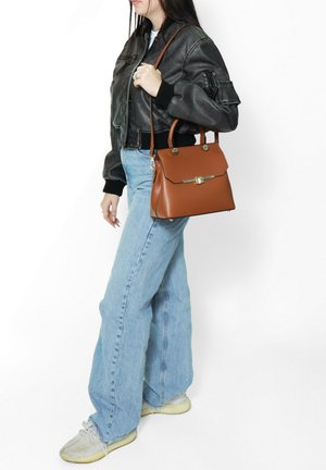 Borsa a tracolla in pelle marrone con hardware dorato, design strutturato, doppi manici e tracolla staccabile; il modello indossa una giacca bomber nera e jeans.