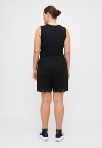 Top nero senza maniche abbinato a pantaloni corti neri con orlo in pizzo a scallop, indossato con sneakers alte nere. Sfondo semplice.