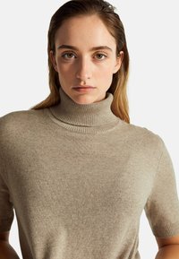 Junge Frau mit hellbraunen Haaren, die einen beigen kurzärmeligen Rollkragenpullover vor einem schlichten hellen Hintergrund trägt.
