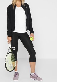 Czarna bomberka na białej koszulce z dekoltem w serek, zestawiona z czarnymi legginsami typu capri z białym logo. Szare buty sportowe i rakieta tenisowa.