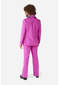 Abito rosa brillante con un colletto di paillettes, caratterizzato da una giacca sartoriale e pantaloni svasati. Il modello ha anche i capelli ricci e scarpe nere.