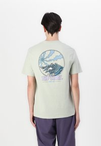 Lys grønn bomull t-skjorte med et sirkulært grafikk på baksiden, med stiliserte bølger og "RIP CURL" tekst i lilla. Korte ermer.