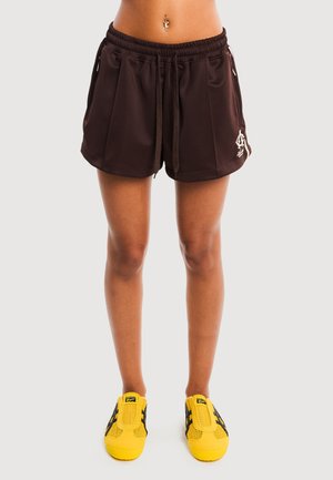Personne portant un short de sport noir et des baskets jaune vif, debout devant un fond uni.