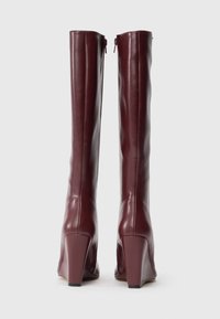 Tall burgundy leather boots with block heels, viewed from the back, standing upright on a white background.