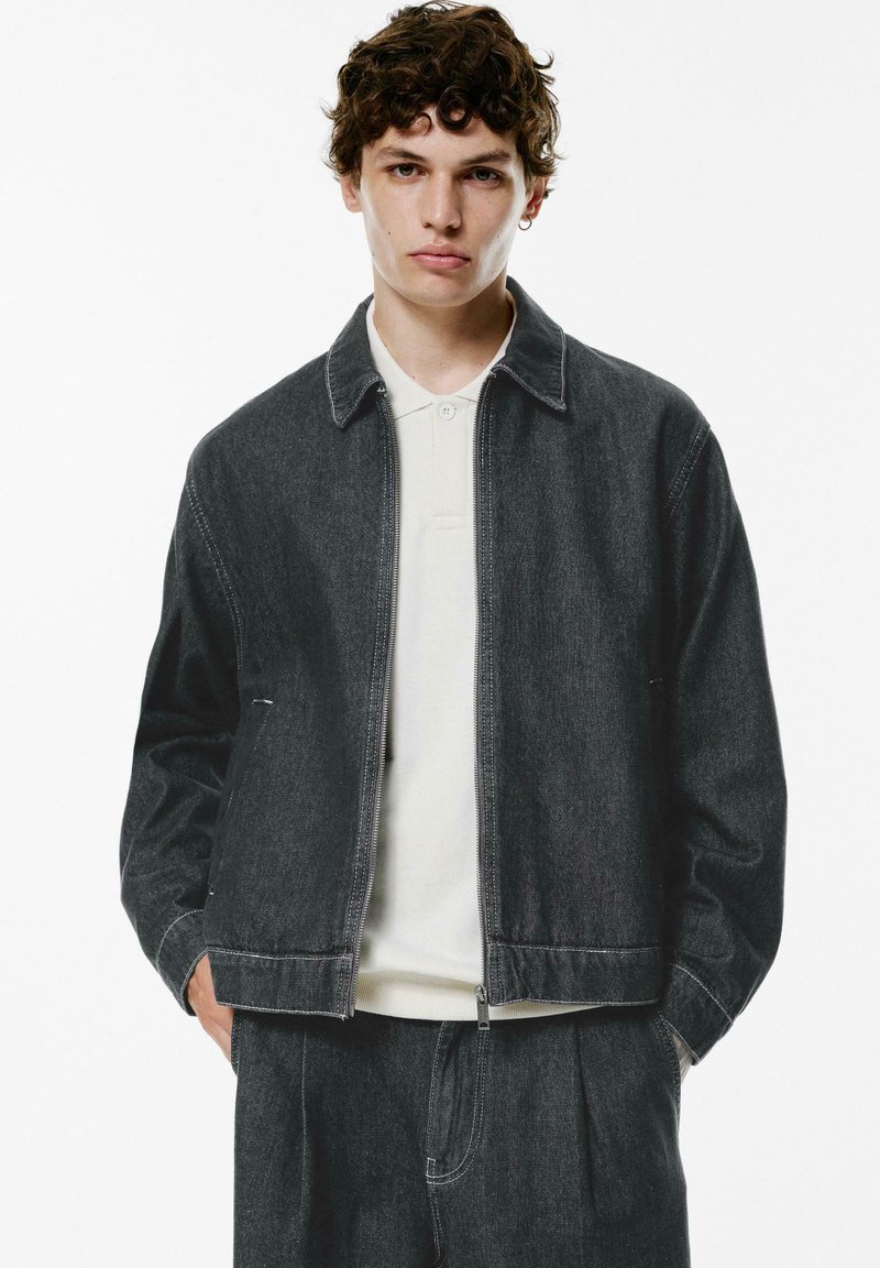 Giubbotto in denim scuro con chiusura a zip, colletto da camicia e tasche laterali. Indossato sopra un polo di colore chiaro, con cuciture a contrasto.
