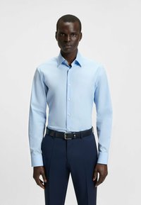 Camicia azzurra a maniche lunghe con colletto classico e chiusura a bottoni; abbinata a pantaloni blu scuro e una cintura nera con fibbia d'argento.