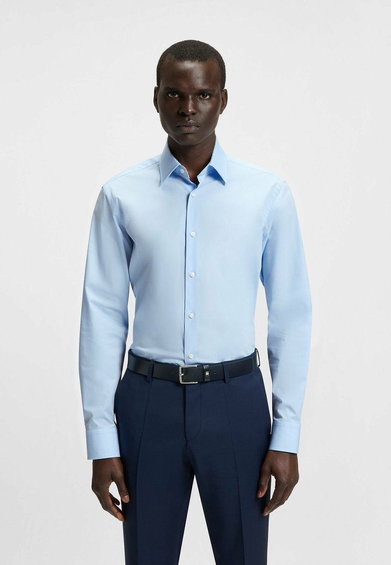 Camicia azzurra a maniche lunghe con colletto classico e chiusura a bottoni; abbinata a pantaloni blu scuro e una cintura nera con fibbia d'argento.