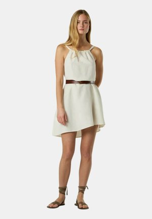 Giovane donna in piedi con un vestito smanicato panna con cintura marrone e sandali bassi marroni con lacci, su uno sfondo chiaro.