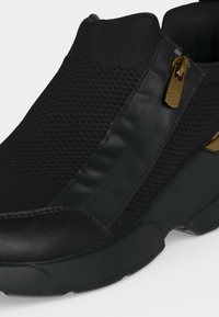 Μαύρο sneaker slip-on με υφασμάτινο πλεκτό επάνω μέρος, ομαλές επιφάνειες και χρυσή λεπτομέρεια με φερμουάρ στο πλάι.