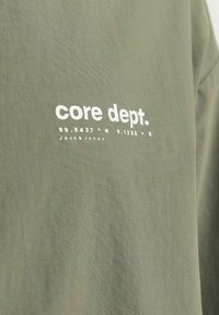 Tamsiai žalias medvilninis marškinėliai su balta užrašu "core dept." ir geografinėmis koordinatėmis. Paprastas, kasdienis dizainas su minkšta tekstūra.